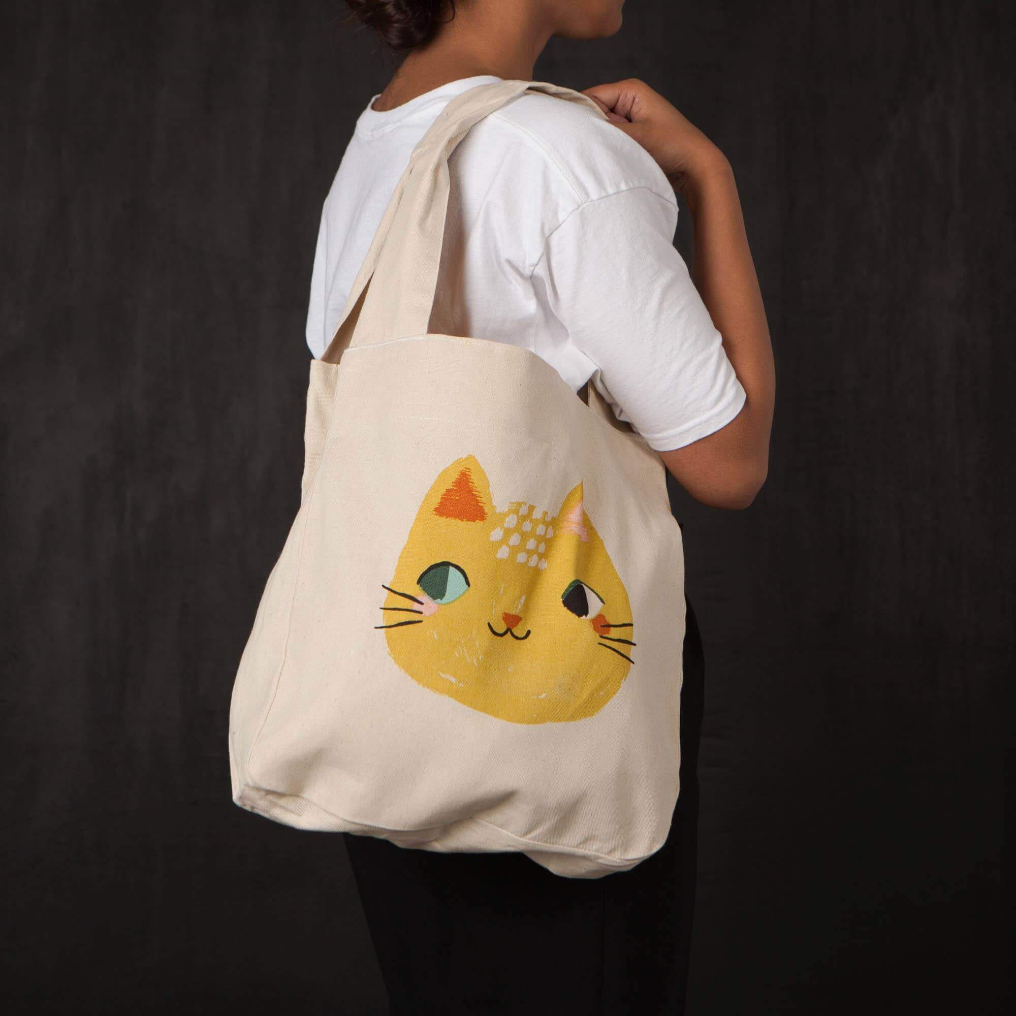 A young woman holds a natural canvas tote bag over her shoulder with a large printed yellow cat face motif on the front