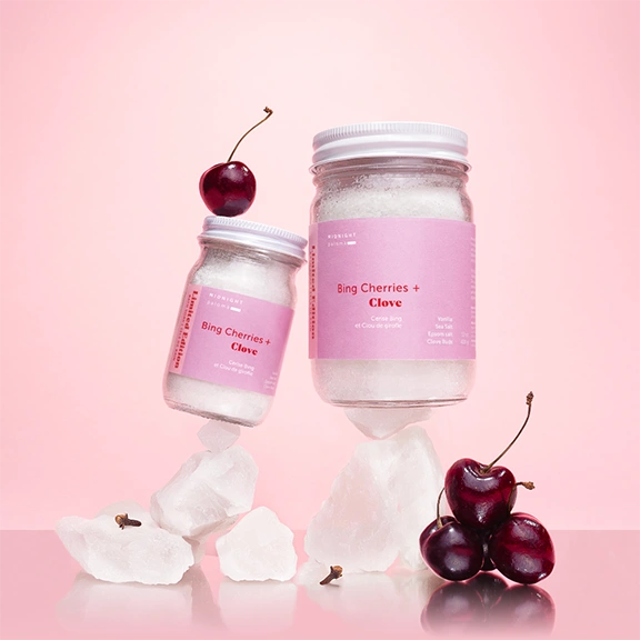 Bing Cherries + Clove Bath Soak jars with cherries on pink background, showcasing luxurious winter bath indulgence.