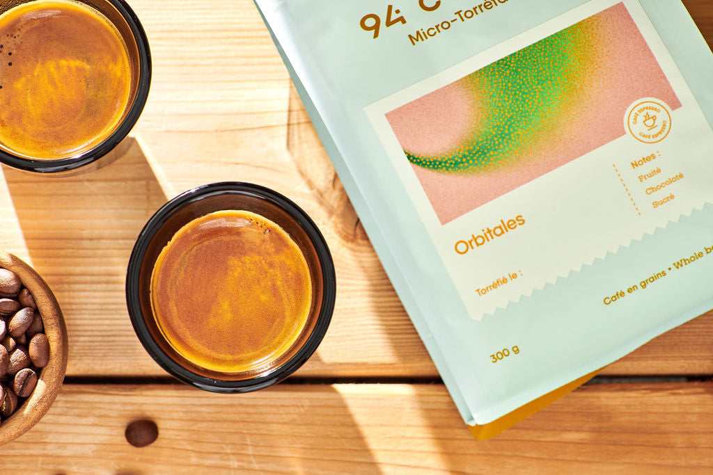 A 300g bag of Orbitales coffee beans by 94 C lies on a light wooden table next to two glasses of espresso and a small bowl of whole coffee beans.
