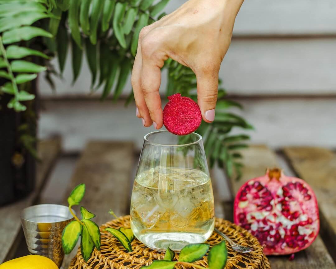 Hand placing a red tea bag into a glass of iced tea on a wooden table with lemons and a pomegranate.