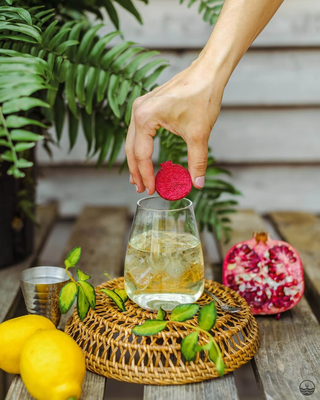 Hand placing a red tea bag into a glass of iced tea on a wooden table with lemons and a pomegranate.