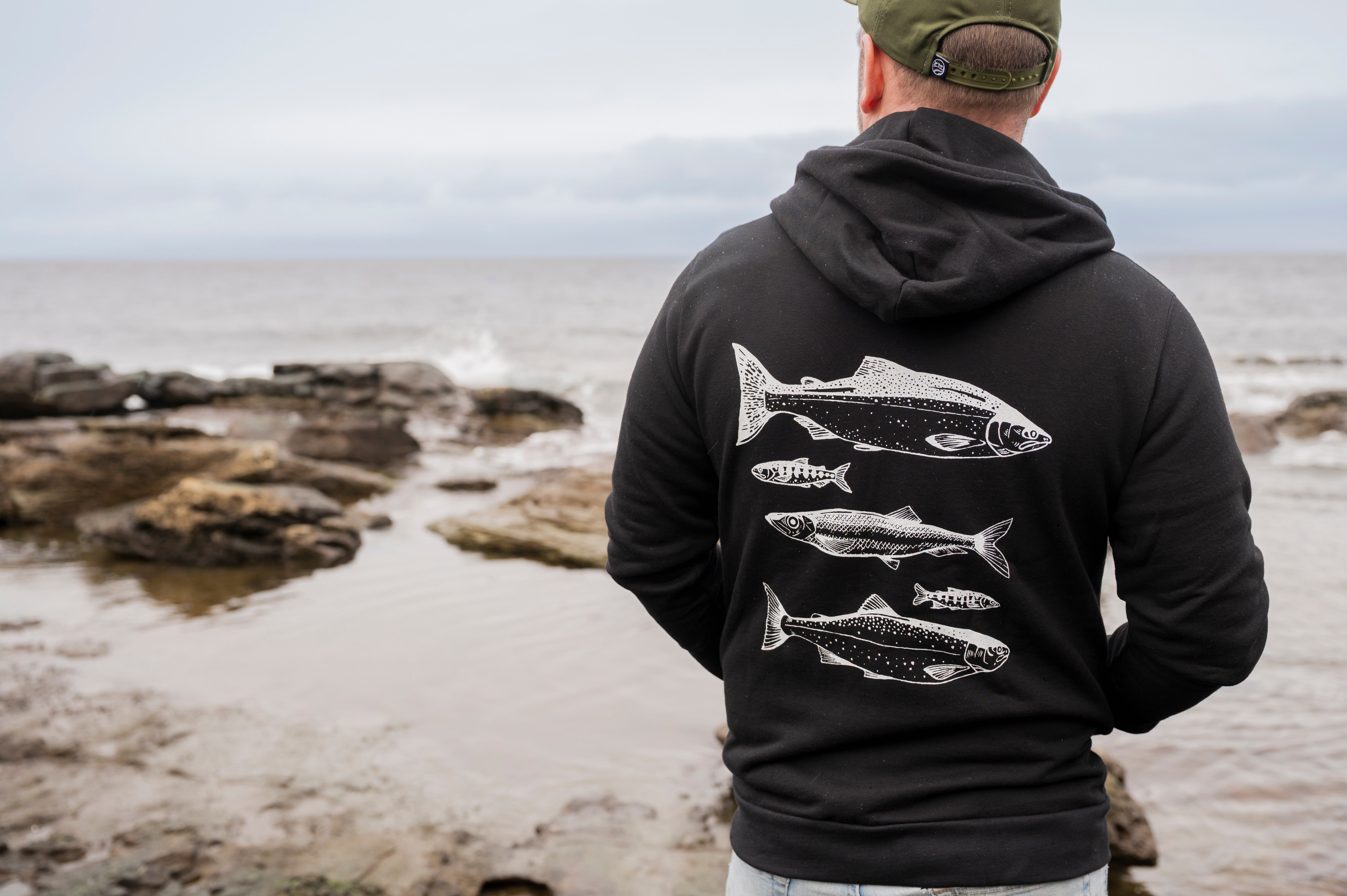 Person wearing a black hoodie with fish design by the ocean
