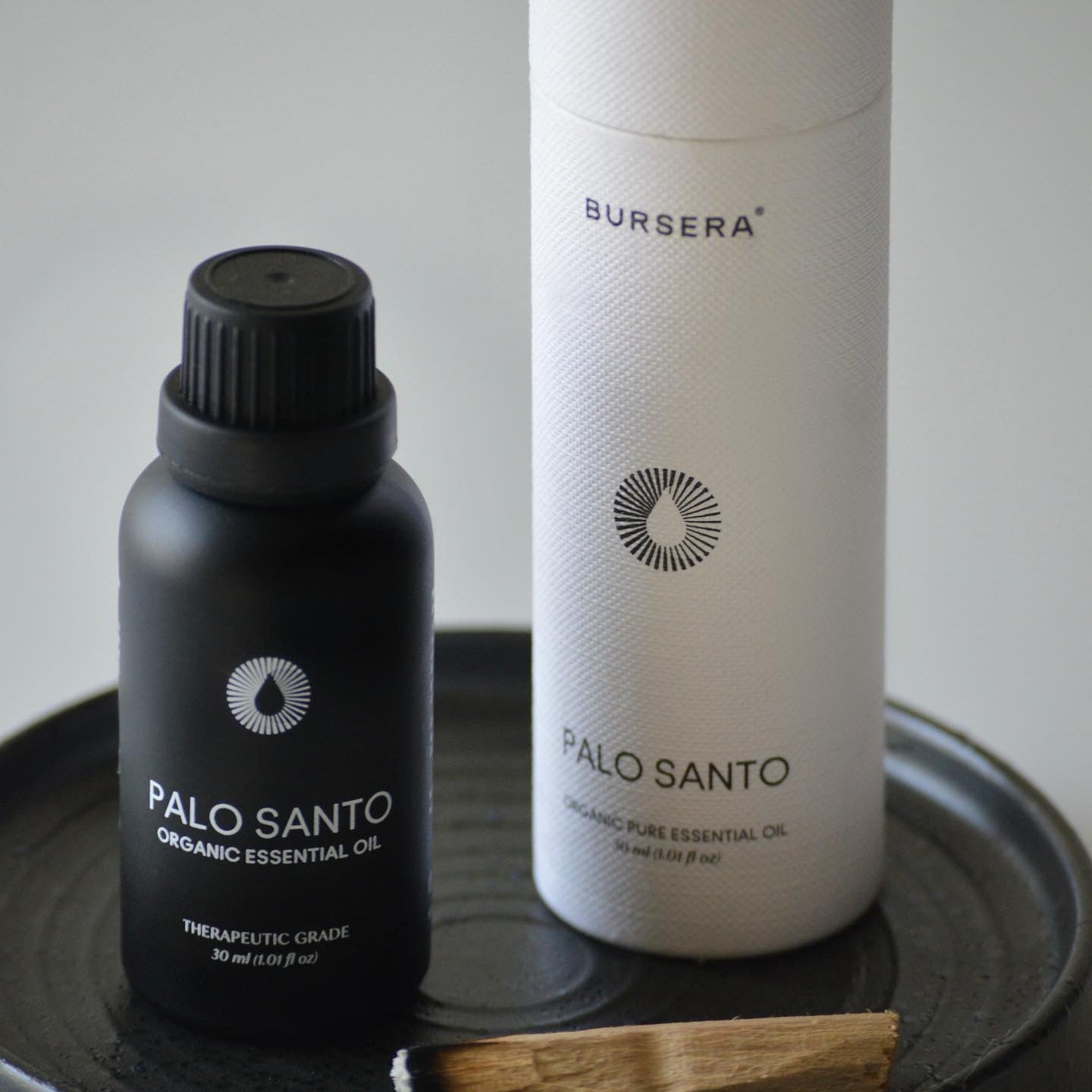 Black bottle and white cylindrical container labeled 'Bursera Palo Santo' on a dark surface.