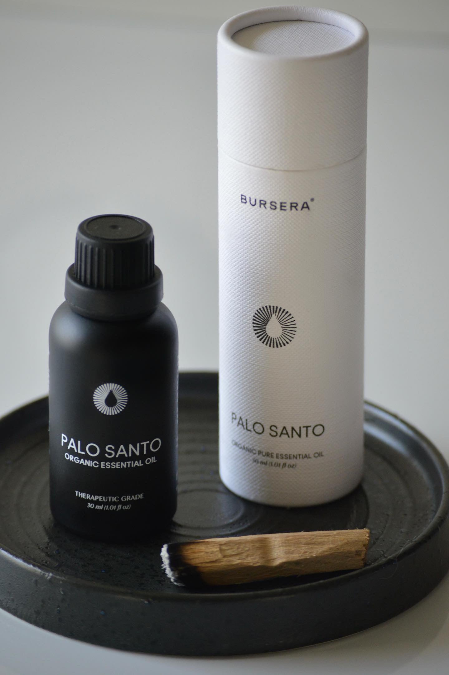 Black bottle and white cylindrical container labeled 'Bursera Palo Santo' on a dark surface.