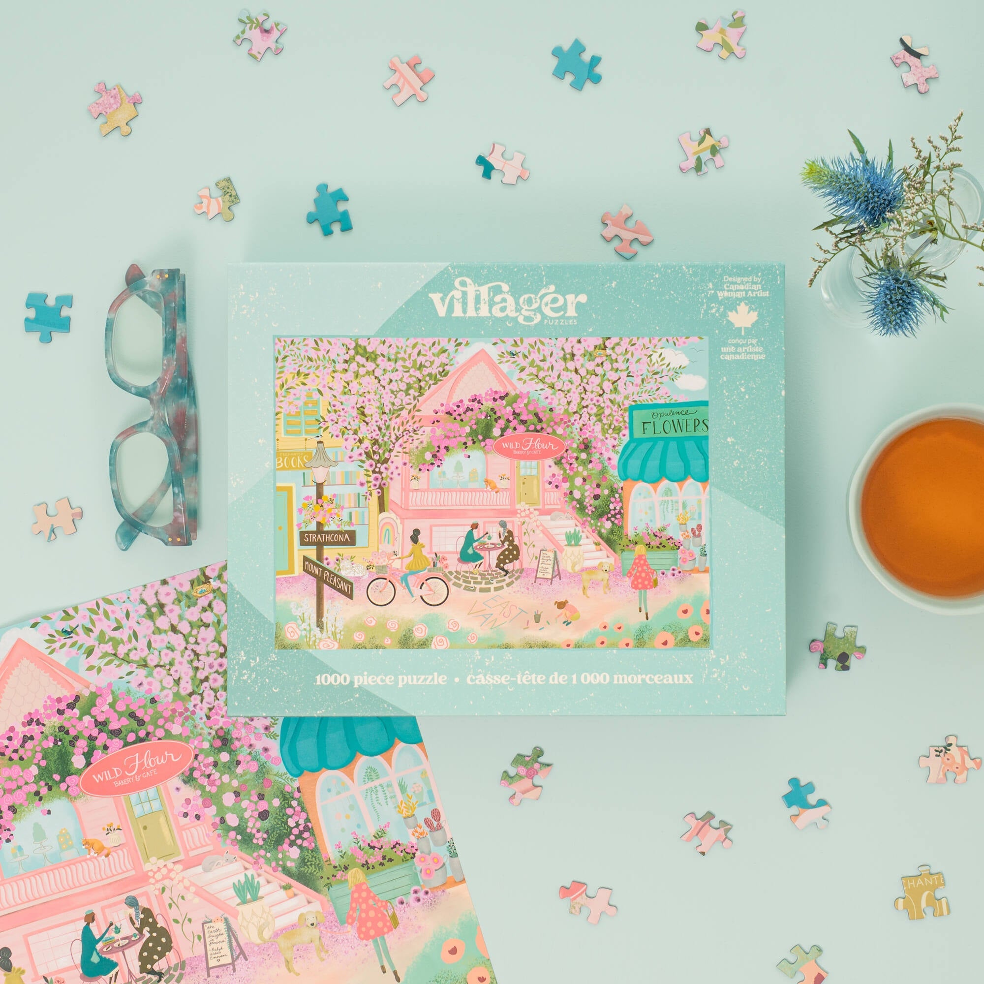 A styled flat-lay of a Villager 1000-piece jigsaw puzzle box on a light blue background, surrounded by loose puzzle pieces, a pair of tortoiseshell glasses, a cup of tea, and floral props. The box features a colorful illustration of a cozy pink flower-shop scene.