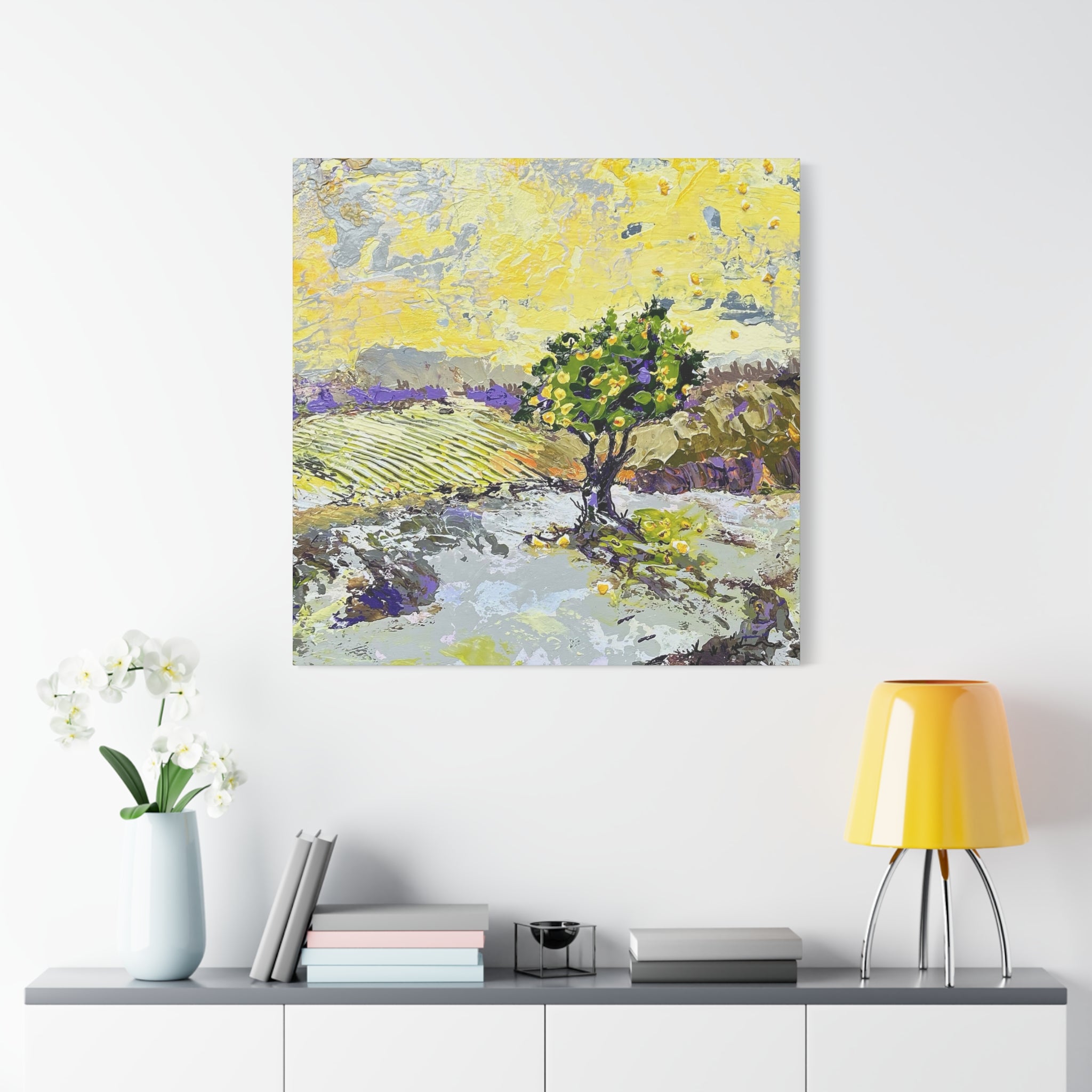 An abstract landscape painting of a tree in a field, displayed on a white wall above a console table with a yellow lamp, books, and a vase of white flowers.