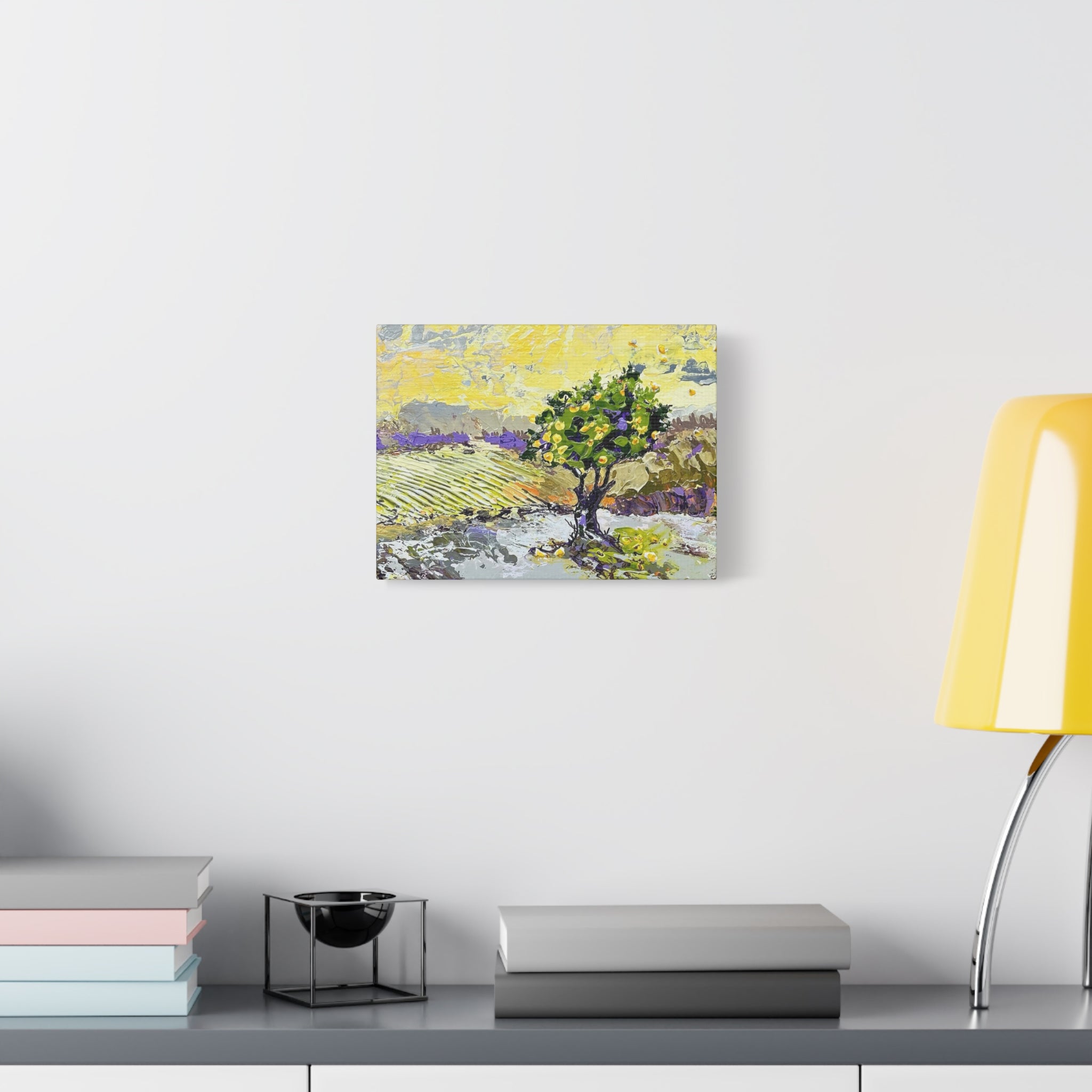 A small square wall art print showing a stylized tree landscape in yellow, green, and purple tones, displayed on a white wall above a shelf with books and a yellow lamp.