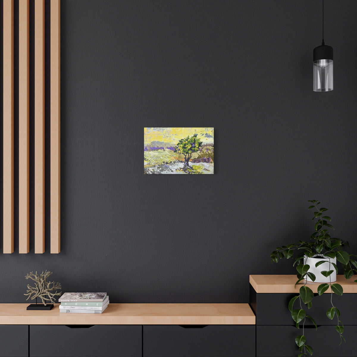 A small colorful landscape art print featuring a tree, displayed on a dark charcoal wall above a light wood console table with books, a plant, and decorative objects.