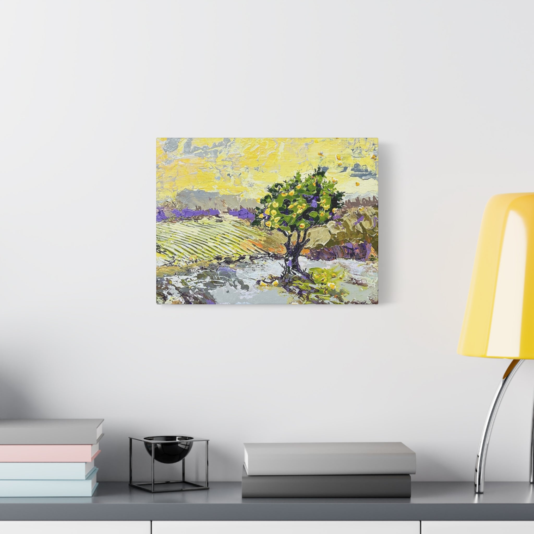 A square canvas art print depicting a stylized landscape with a green tree, yellow sky, and purple fields, displayed on a white wall above a gray console table with books and a yellow lamp.