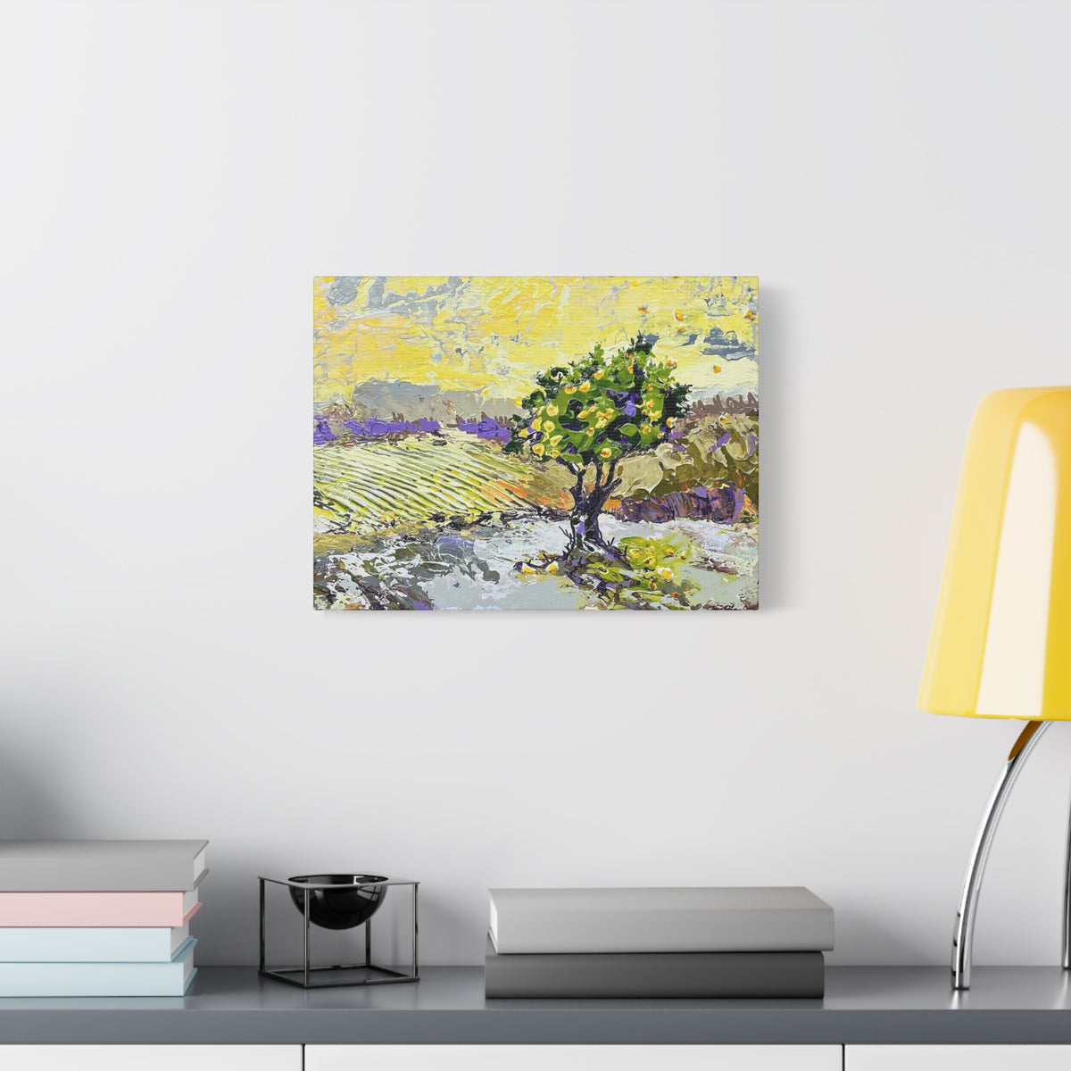 A square canvas art print depicting a stylized landscape with a green tree, yellow sky, and purple fields, displayed on a white wall above a gray console table with books and a yellow lamp.