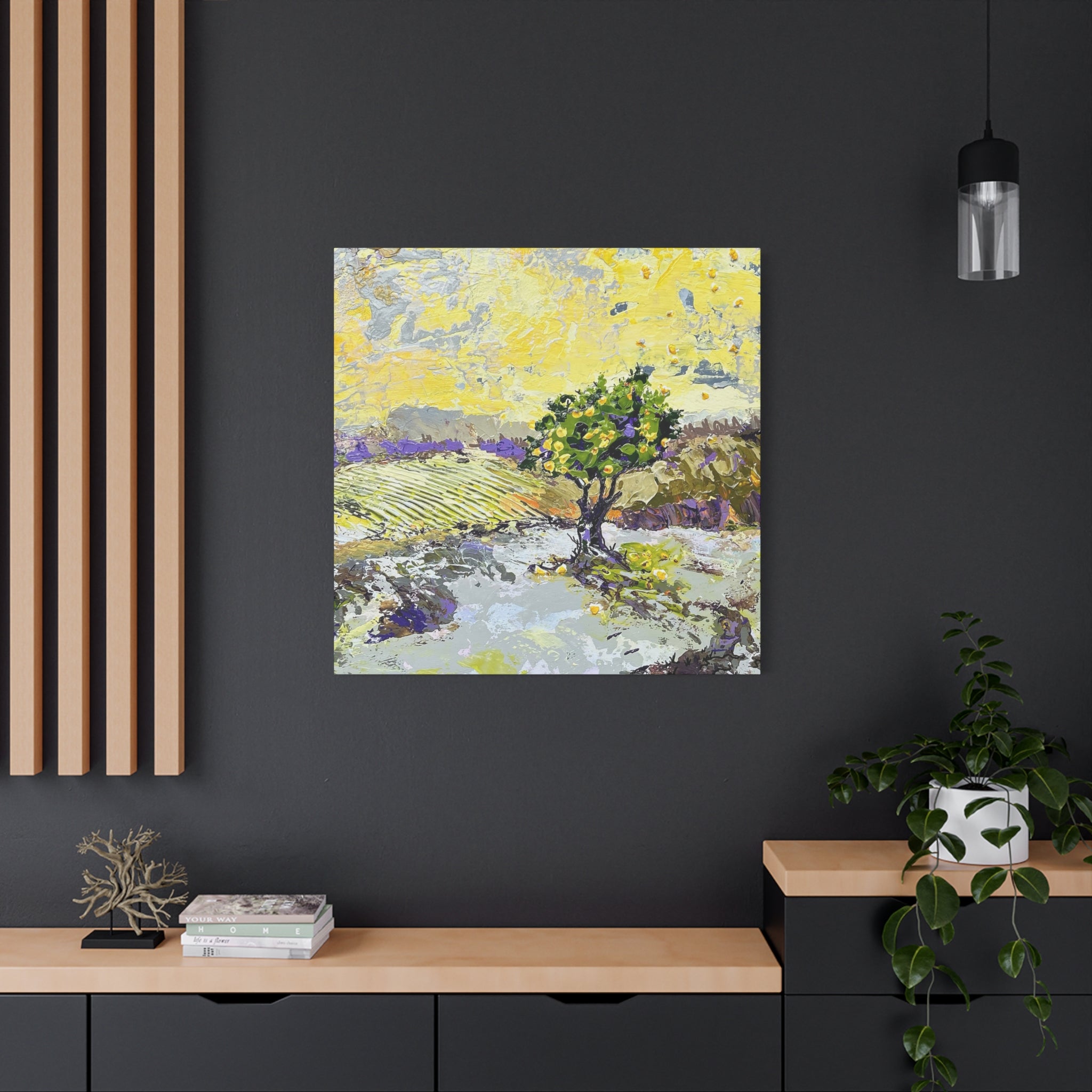 A square canvas painting of a vineyard landscape with a central green tree, displayed on a dark gray wall in a modern room setting with a wooden console table, a potted plant, and a hanging light fixture.