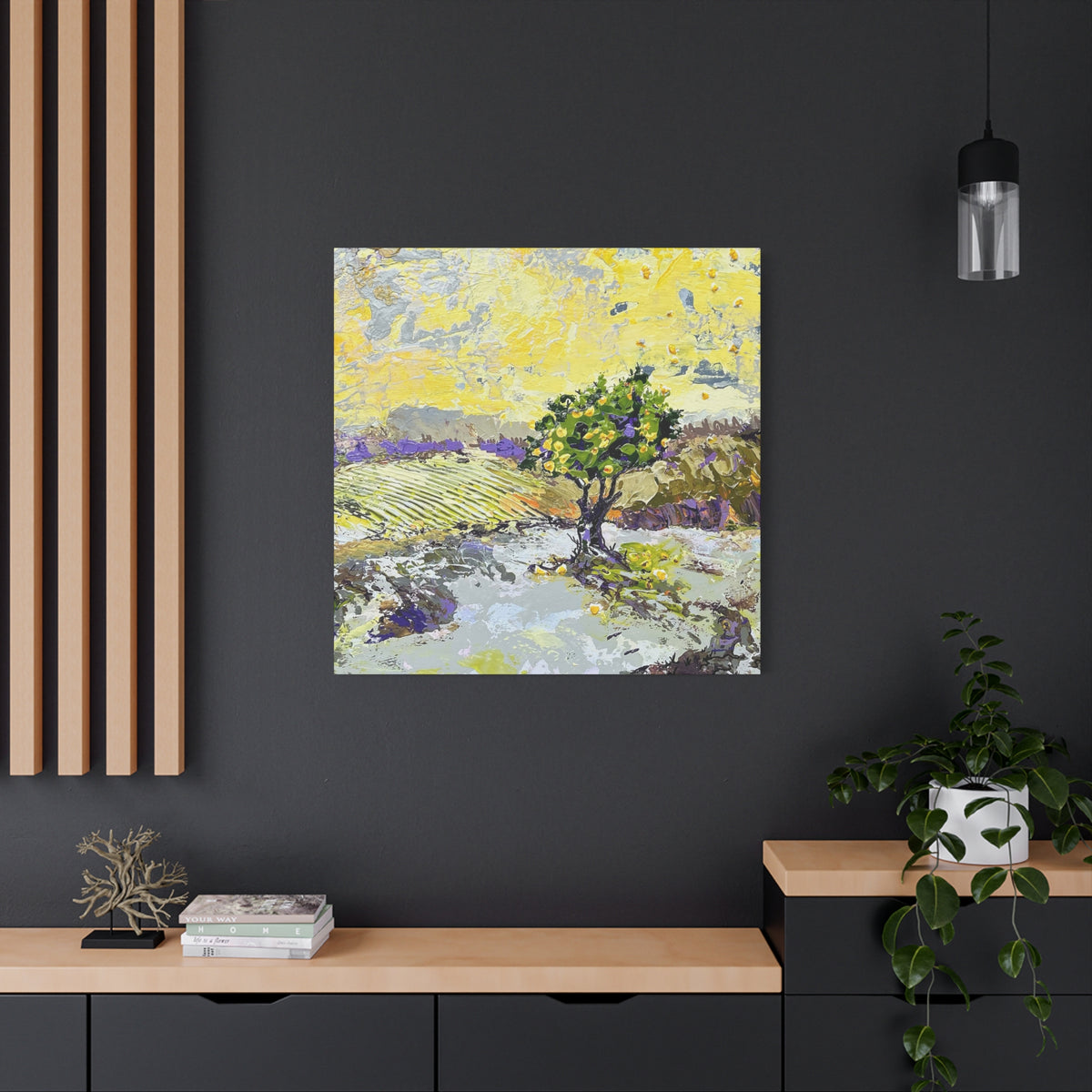 A square canvas painting of a vineyard landscape with a central green tree, displayed on a dark gray wall in a modern room setting with a wooden console table, a potted plant, and a hanging light fixture.