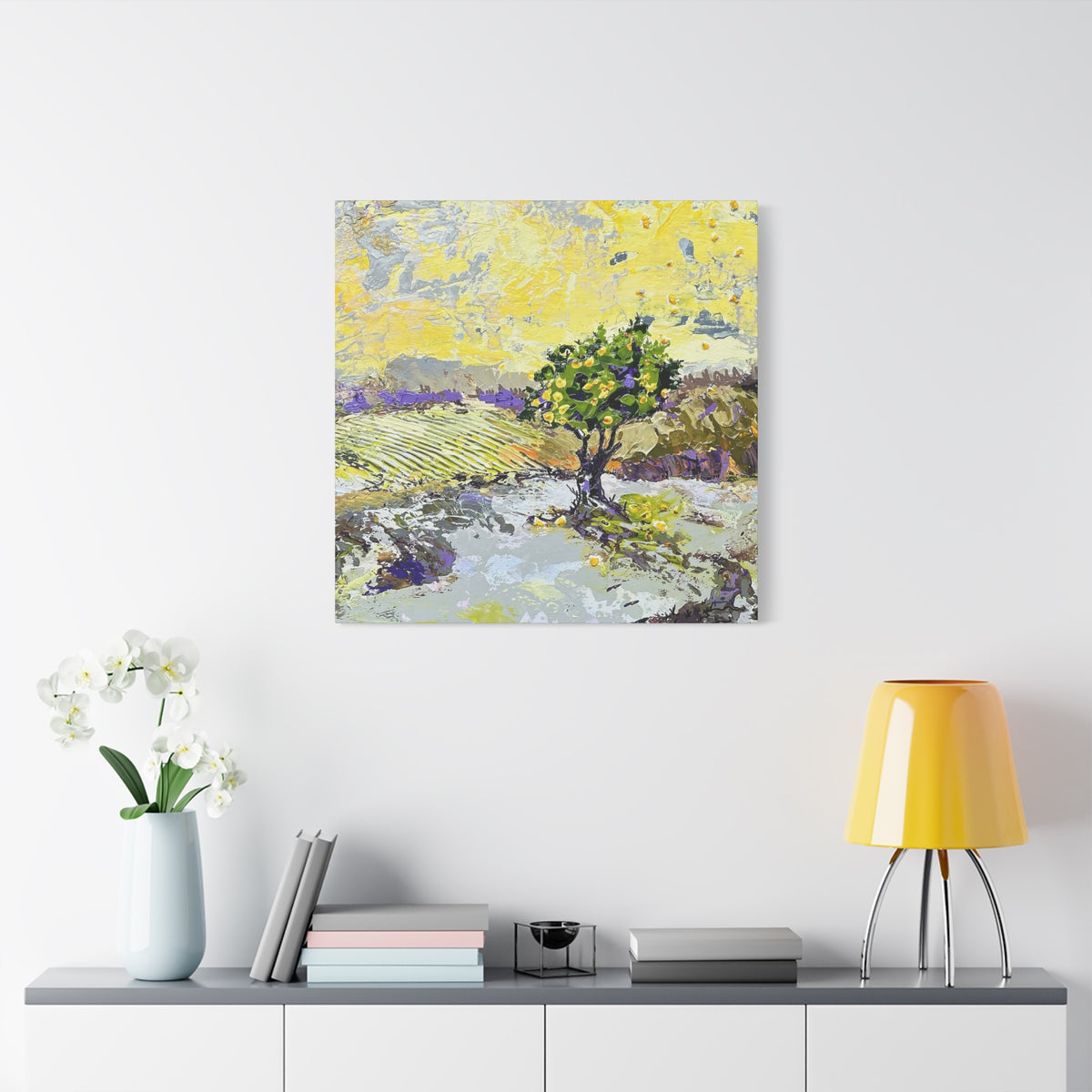 A square canvas art print featuring an impressionistic landscape painting with a green tree, yellow sky, and purple fields, displayed on a white wall above a console table with a vase of white flowers, books, and a yellow lamp.