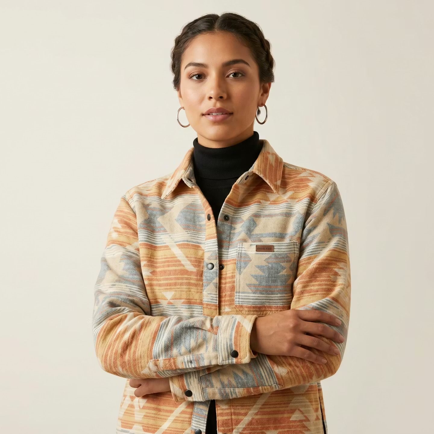 A woman wearing a long-sleeve button-up shirt with a southwestern geometric pattern in orange, beige, and blue, layered over a black turtleneck, against a plain light beige background.