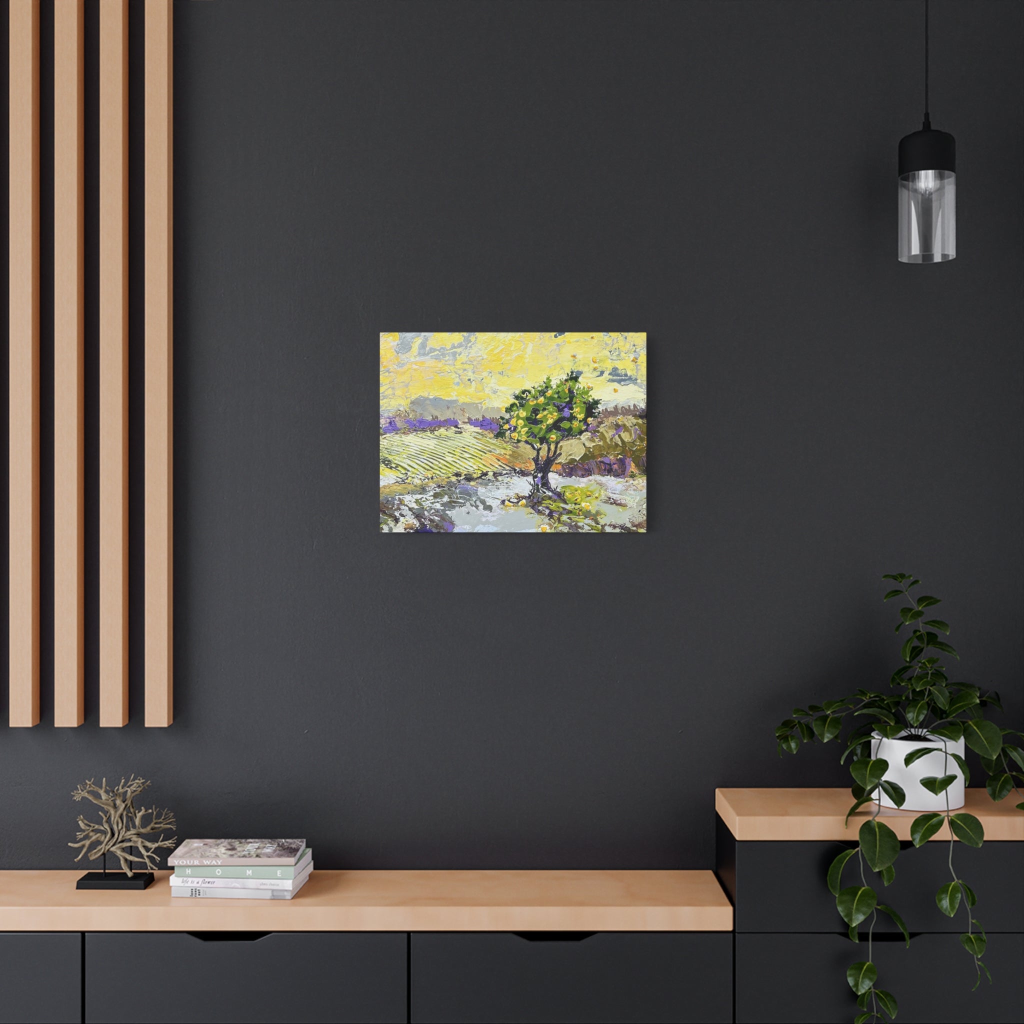 A small square landscape painting hanging on a dark charcoal wall above a light wood console table. The artwork depicts a tree and pale yellow field with purple and green accents.