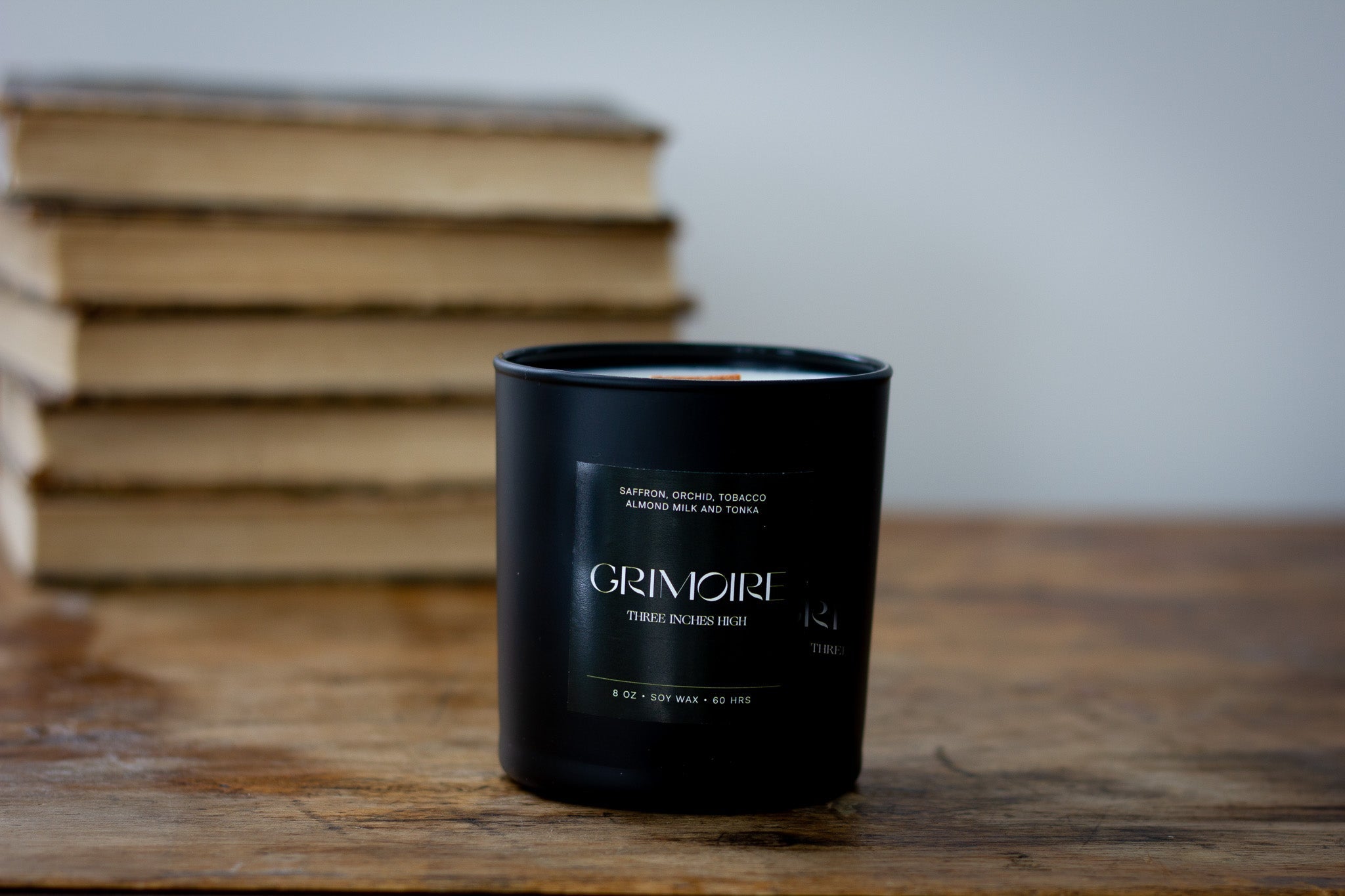 Black candle with 'GRIMOIR' branding on a wooden surface with books in the background