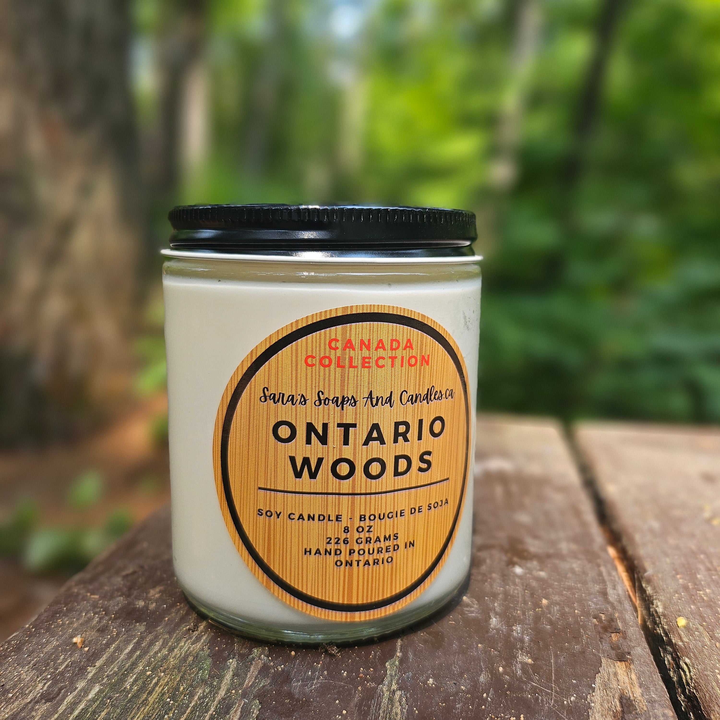 Candle labeled 'Ontario Woods' from the Canada Collection on a wooden surface with a forest background.