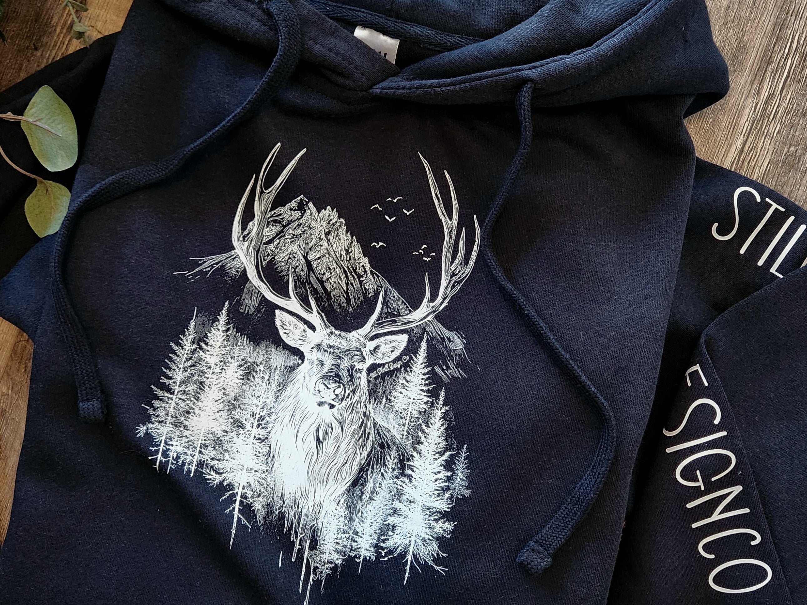 Still North Design Co Navy blue hoodie with deer design on a wooden surface