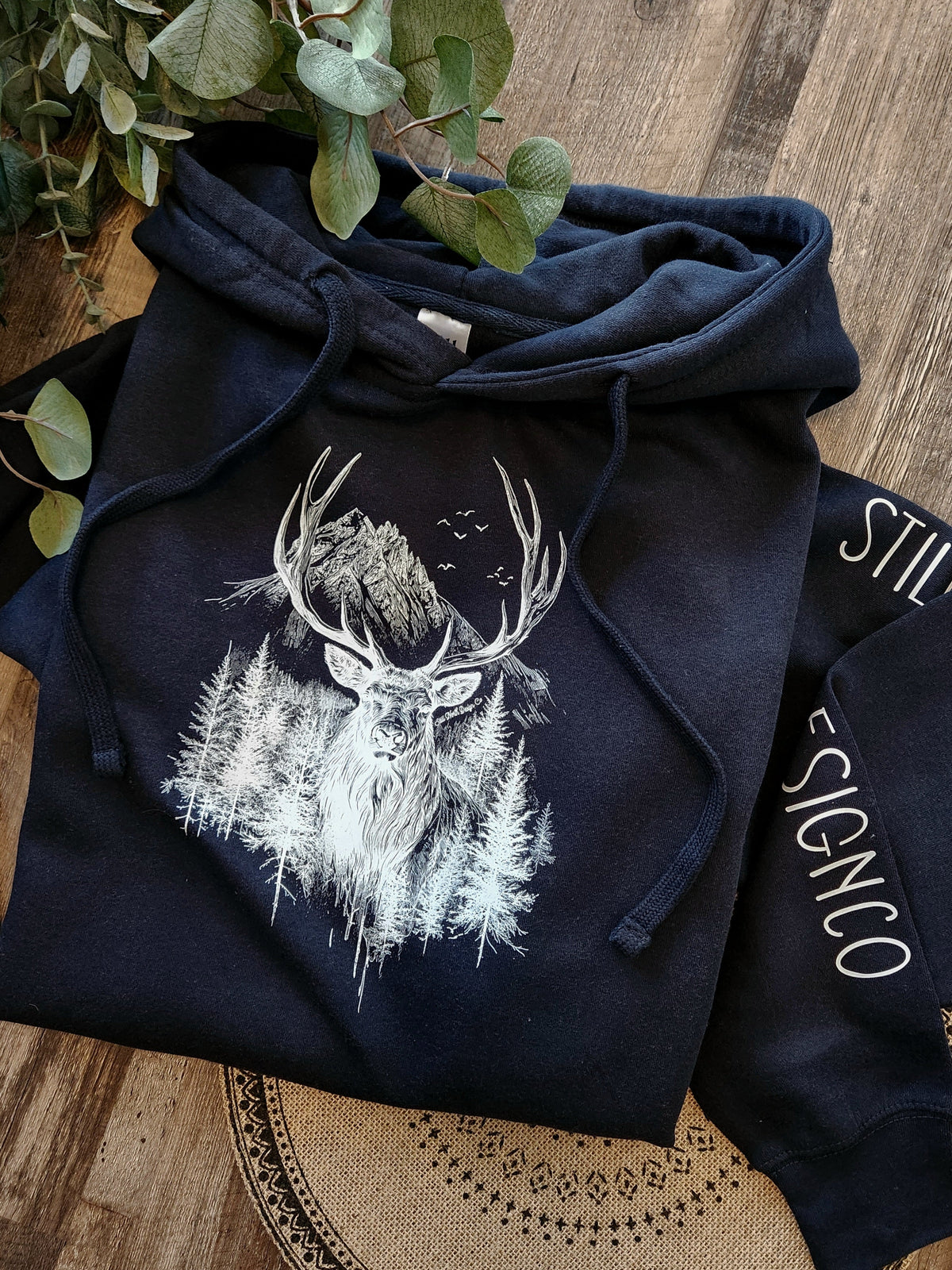Still North Design Co Navy blue hoodie with deer design on a wooden surface