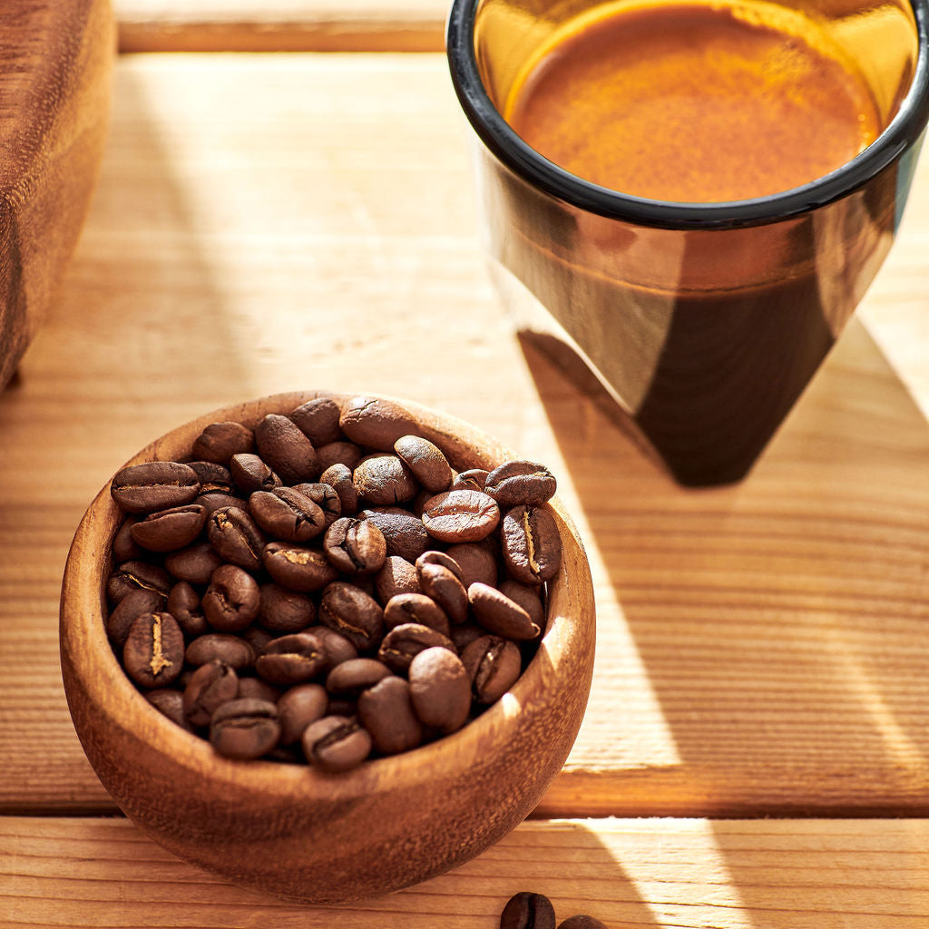 A wooden bowl filled with roasted coffee beans sits on a light wood table beside a dark amber glass cup containing espresso, with a few beans scattered nearby in warm natural light.