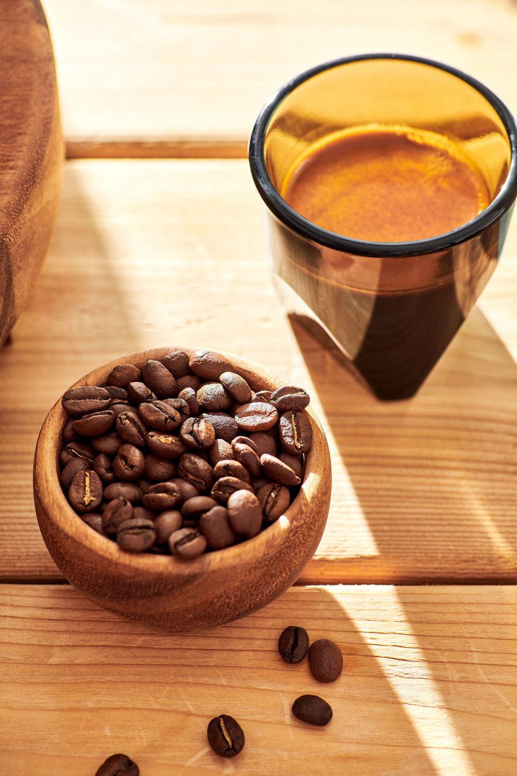 A wooden bowl filled with roasted coffee beans sits on a light wood table beside a dark amber glass cup containing espresso, with a few beans scattered nearby in warm natural light.