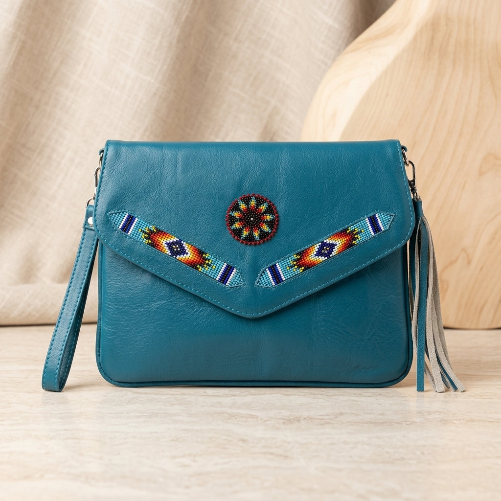 OldTribes A teal blue leather crossbody bag with a flap closure, decorated with a circular beaded medallion and geometric beaded accents on the flap. The bag features a matching wristlet strap, tassel detail, and is displayed on a light wood surface with a neutral fabric backdrop.