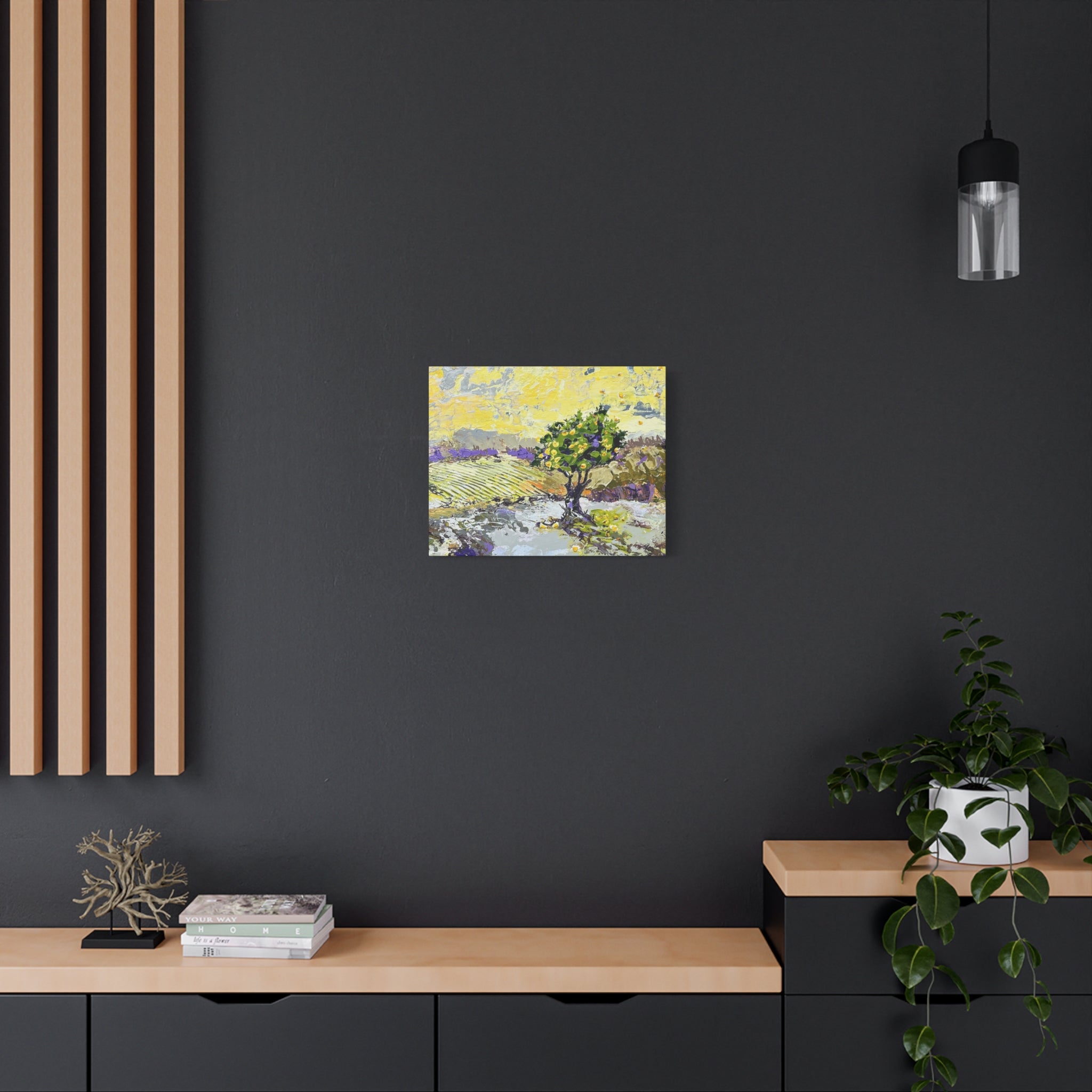 A small square canvas painting of a tree in a landscape hangs on a dark charcoal wall above a light wood console table in a modern room setting.