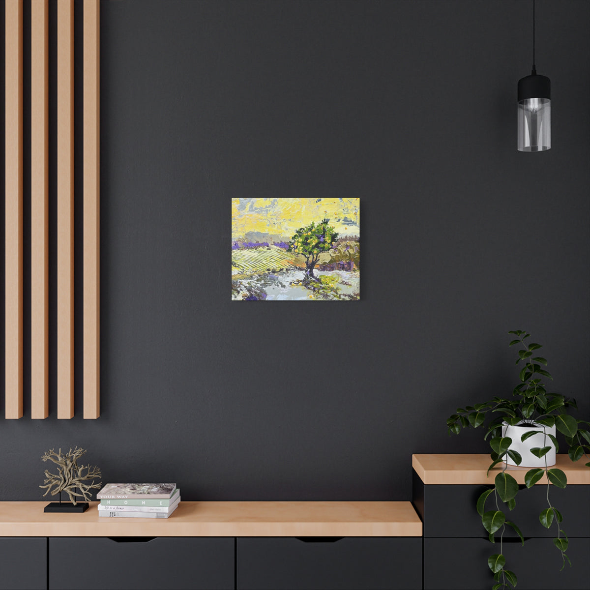 A small square canvas painting of a tree in a landscape hangs on a dark charcoal wall above a light wood console table in a modern room setting.