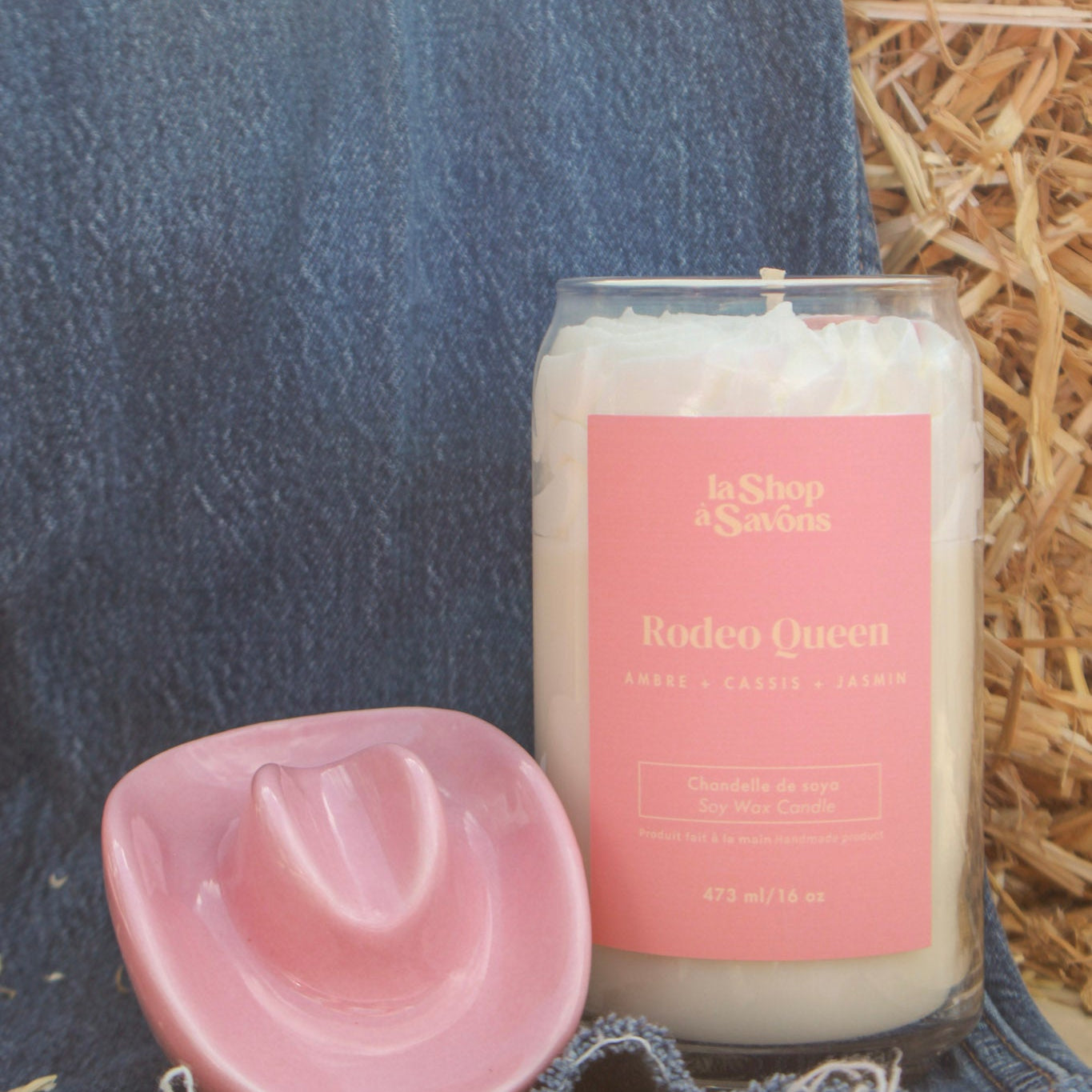 Candle labeled 'Rodeo Queen' with a pink cowboy hat on hay and denim background