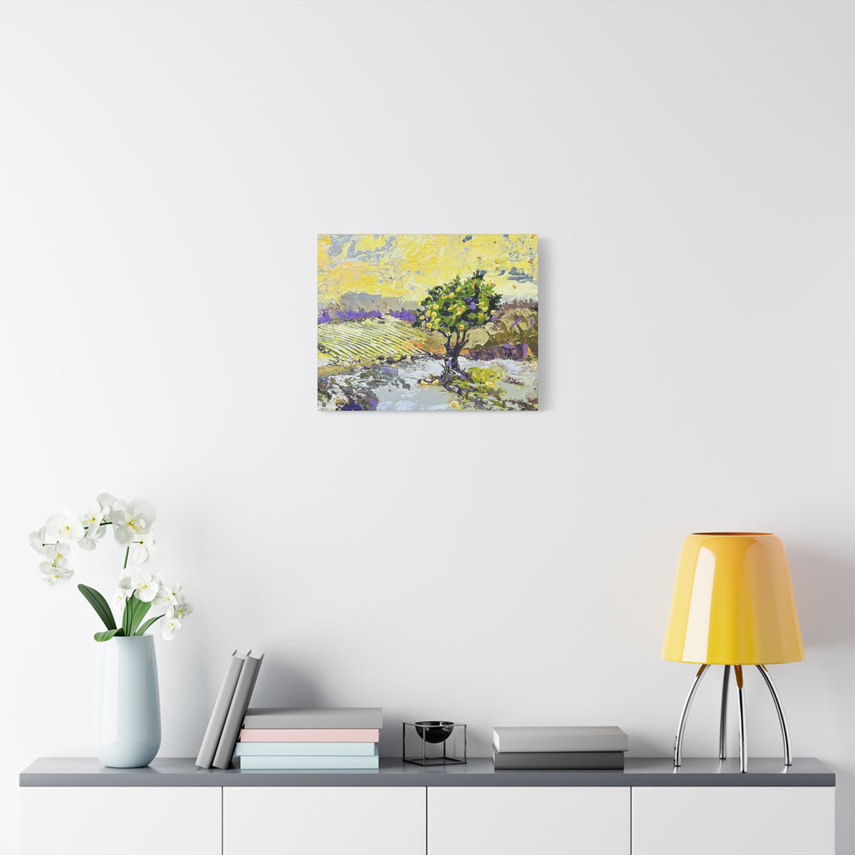 A square canvas art print depicting a stylized landscape with a green tree and purple fields, displayed on a white wall above a gray console table styled with a vase of white flowers, books, and a yellow lamp.