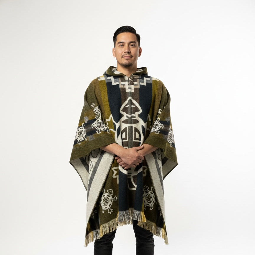 A man wearing a hooded knit poncho with a dark navy blue, olive green, and cream geometric pattern, standing against a plain white studio background.