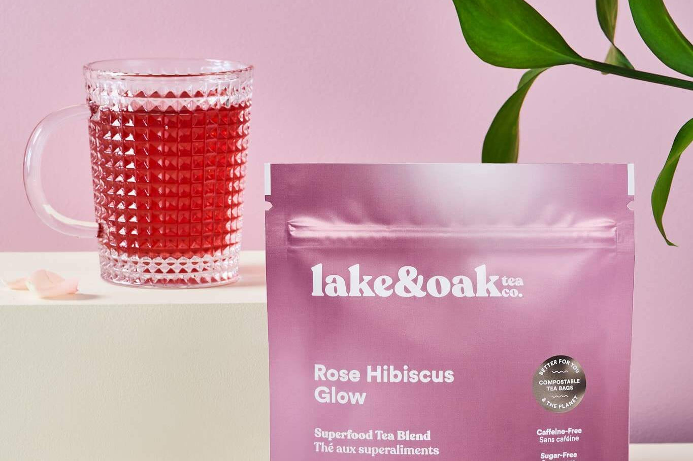 Rose Hibiscus Glow superfood tea package beside a glass of ruby red tea and garnishes on a pastel background.