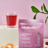 Rose Hibiscus Glow superfood tea package beside a glass of ruby red tea and garnishes on a pastel background.