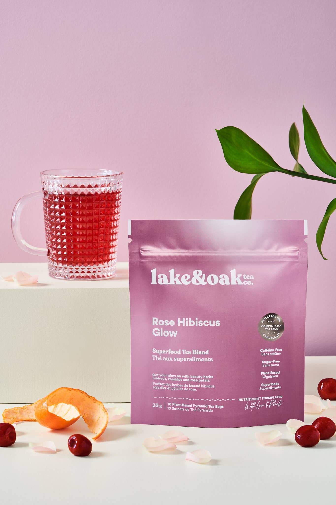 Rose Hibiscus Glow superfood tea package beside a glass of ruby red tea and garnishes on a pastel background.