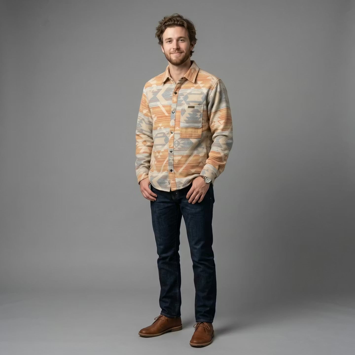 A man modeling a long-sleeve button-up shirt with a warm-toned abstract pattern in orange, beige, and gray, paired with dark blue jeans and brown shoes, against a solid gray studio background.