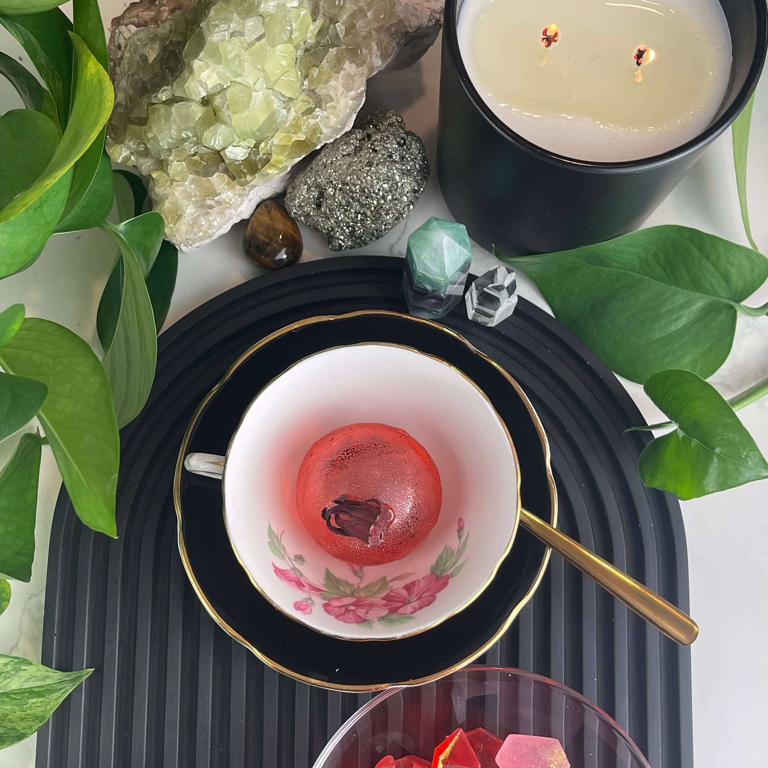 Berry Hibiscus Tea Bomb served in a vintage cup, alongside colorful treats and a candle, surrounded by lush greenery.