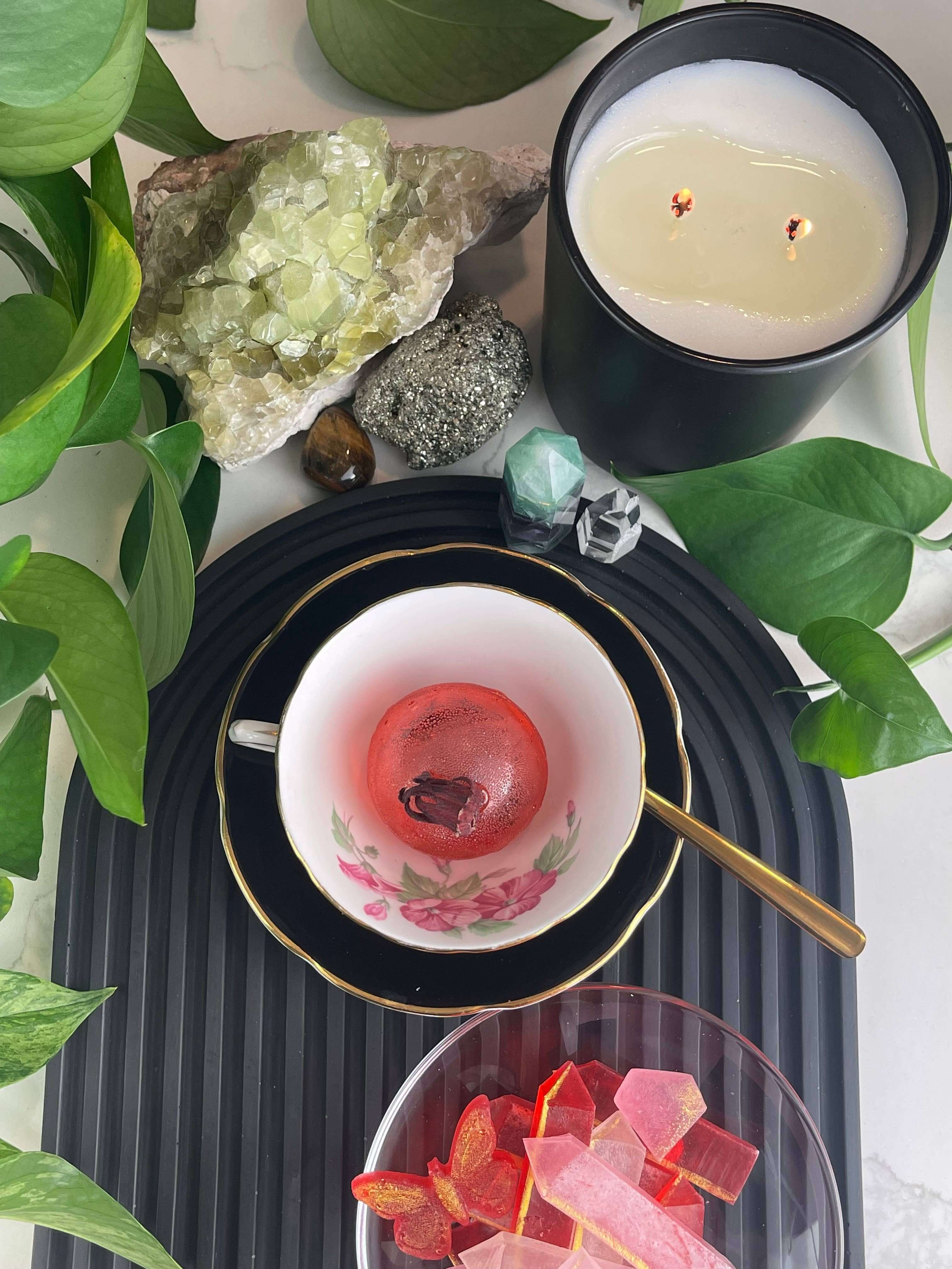 Berry Hibiscus Tea Bomb served in a vintage cup, alongside colorful treats and a candle, surrounded by lush greenery.