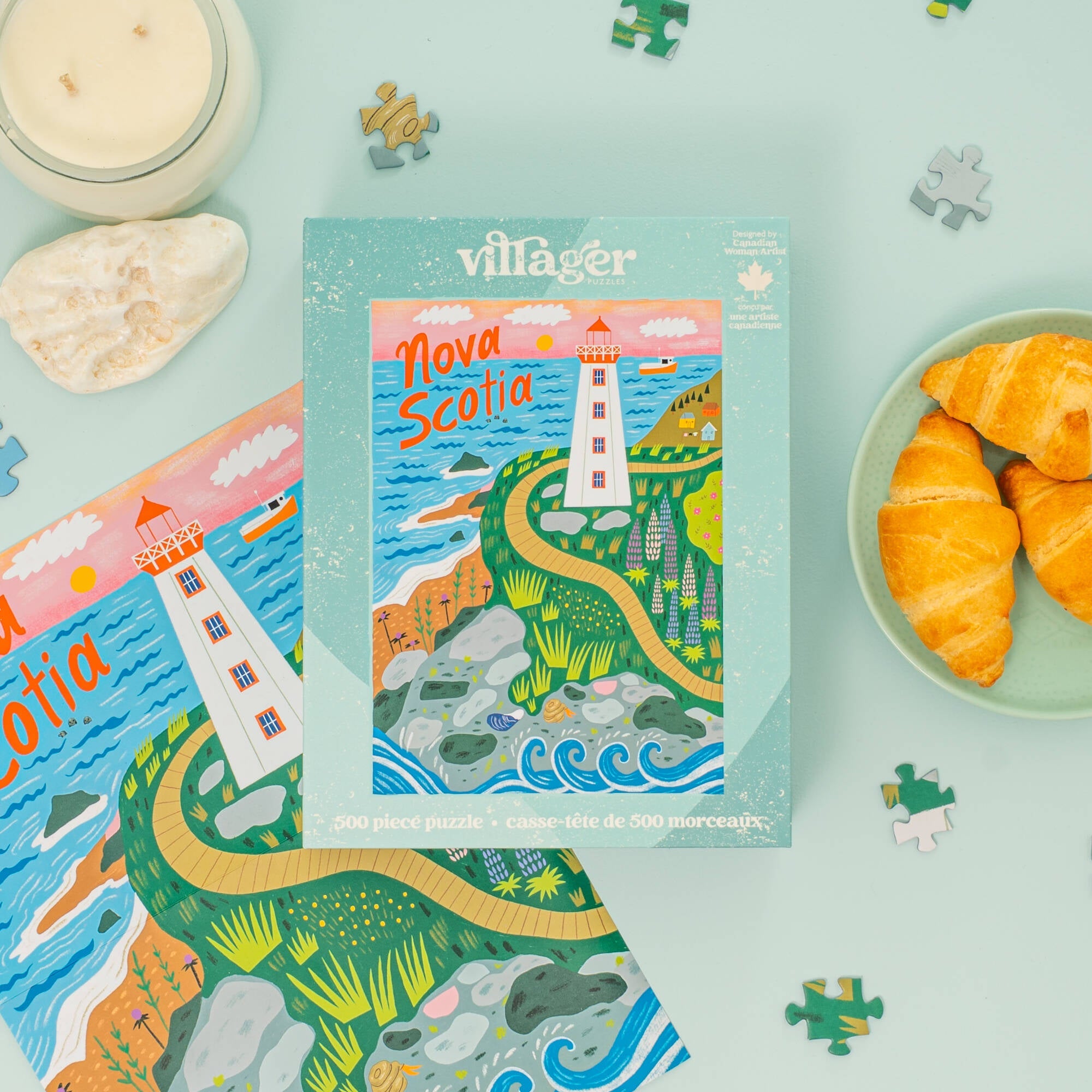 Villager Nova Scotia 500-piece jigsaw puzzle box displayed on a light blue surface, surrounded by the puzzle pieces, a lit candle, a shell, and croissants on a plate.