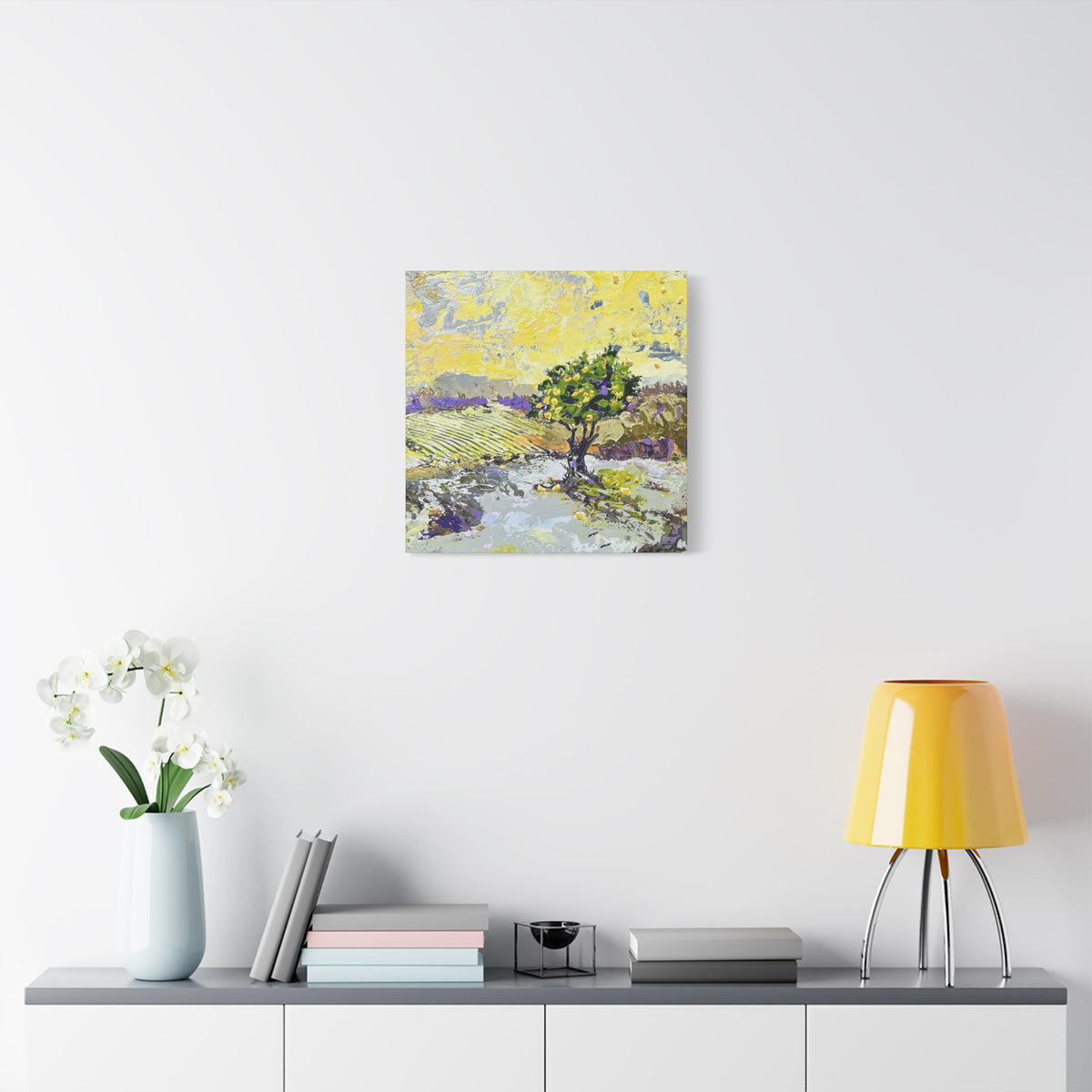 A square canvas art print depicting a stylized landscape with a green tree, yellow sky, and purple fields, displayed on a white wall above a console table with a yellow lamp, books, and a vase of white flowers.