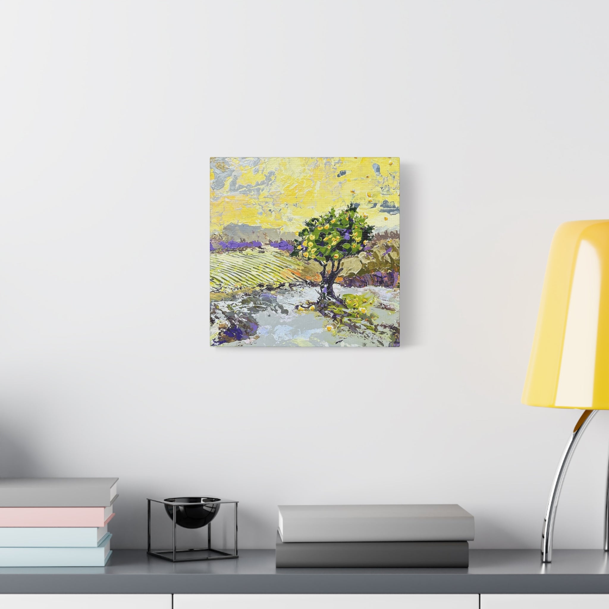 A square canvas art print featuring an impressionistic landscape painting of a green tree in a yellow field, displayed on a white wall above a gray shelf with books and a yellow lamp.