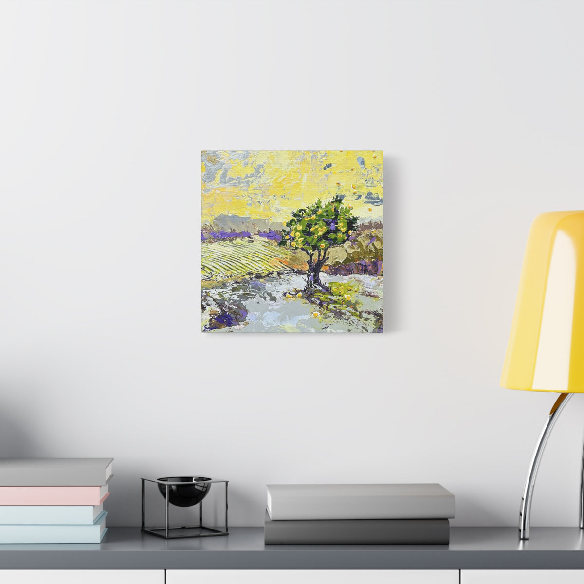 A square canvas art print featuring an impressionistic landscape painting of a green tree in a yellow field, displayed on a white wall above a gray shelf with books and a yellow lamp.