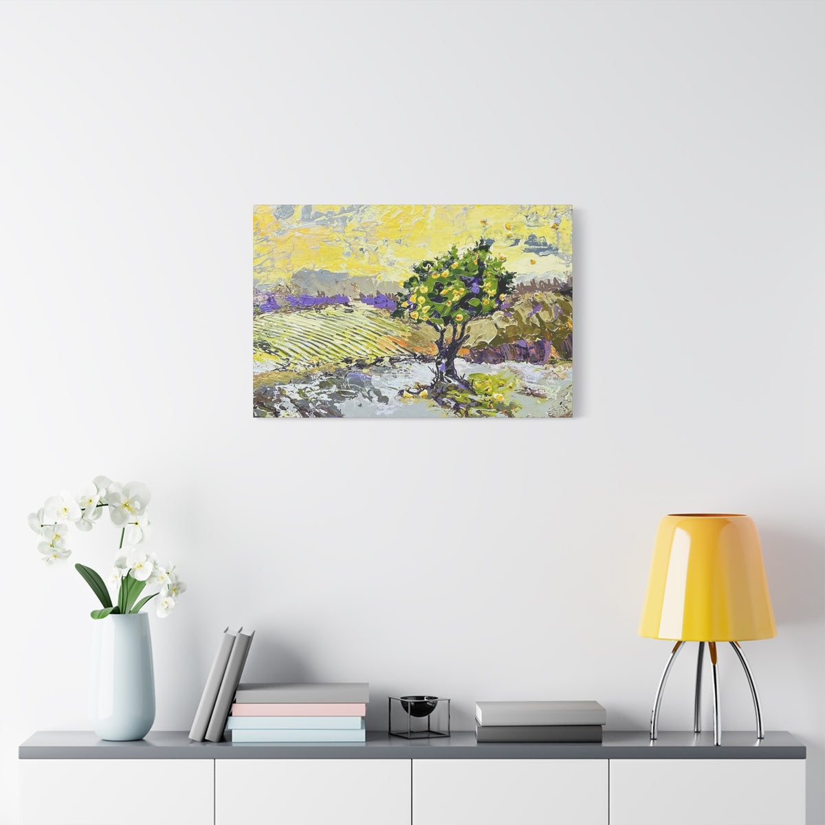 A square canvas art print depicting a stylized landscape with a green tree and yellow fields, displayed on a white wall above a console table with a vase of white flowers, books, and a yellow lamp.