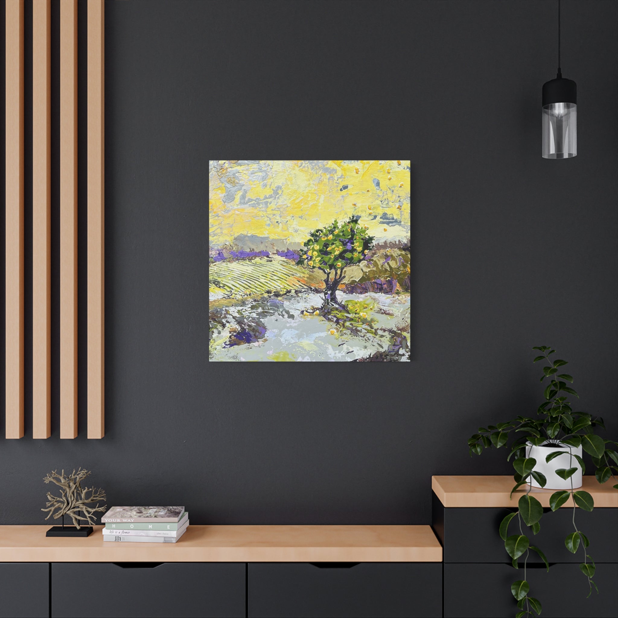 A square canvas art print featuring an impressionistic landscape painting of a tree in a field, displayed on a dark gray wall in a modern styled room with a wooden console, a potted plant, and a hanging light.