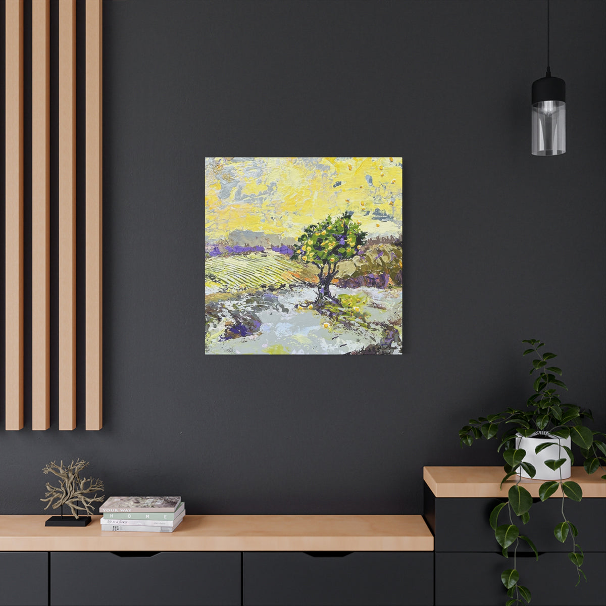 A square canvas art print featuring an impressionistic landscape painting of a tree in a field, displayed on a dark gray wall in a modern styled room with a wooden console, a potted plant, and a hanging light.