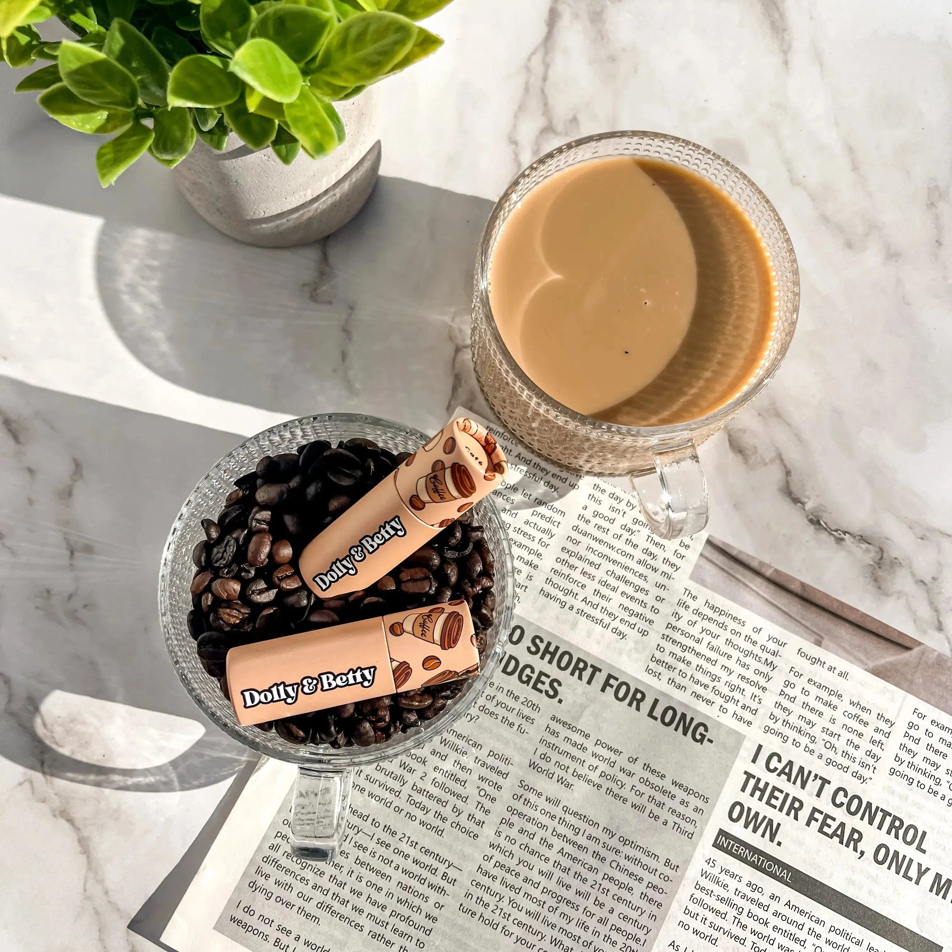 Glass mug with coffee, coffee beans, and Dolly & Betty products on a marble surface with a plant and newspaper.