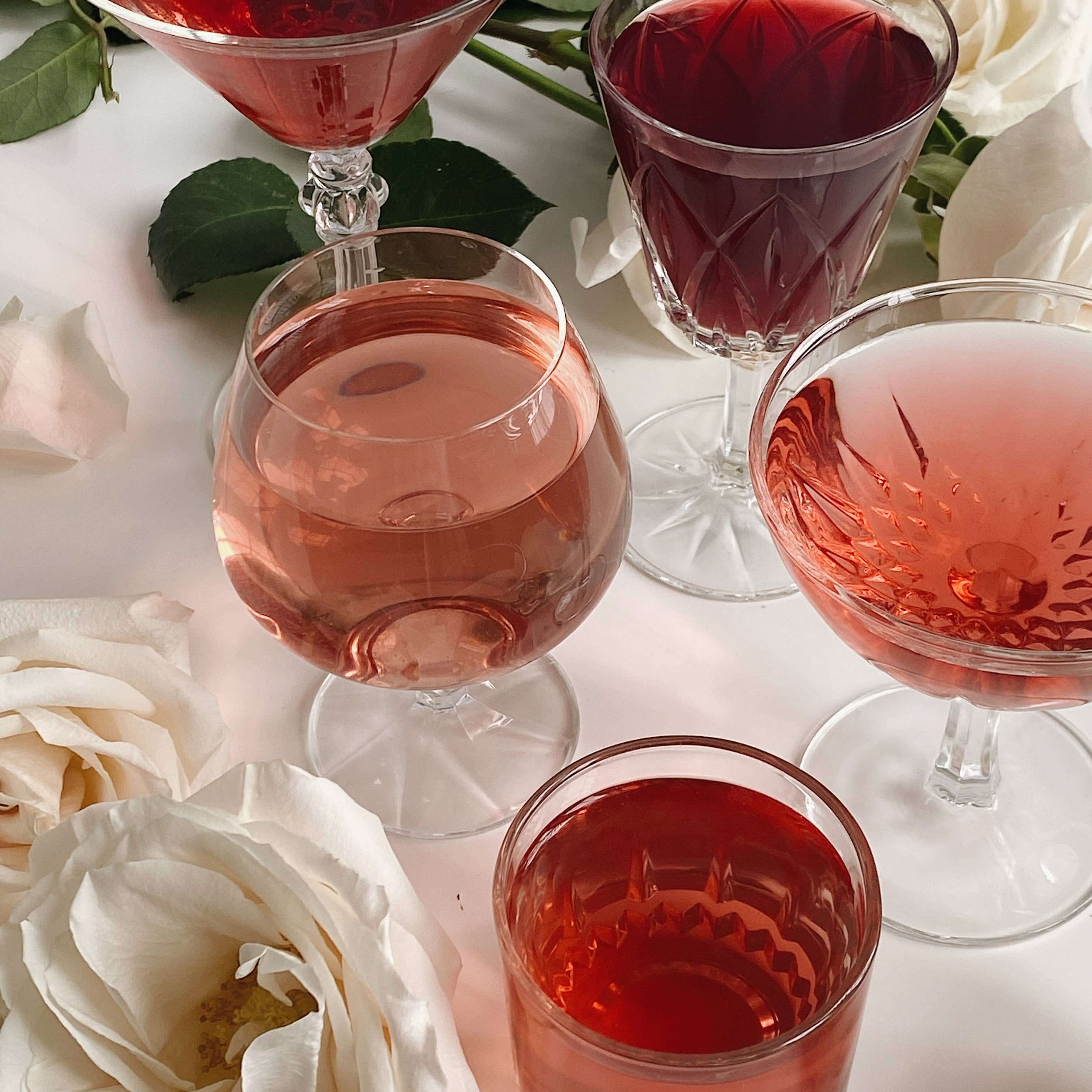 Assorted glasses of vibrant hibiscus tea drinks with white roses on a table, showcasing rose and fruity colors.