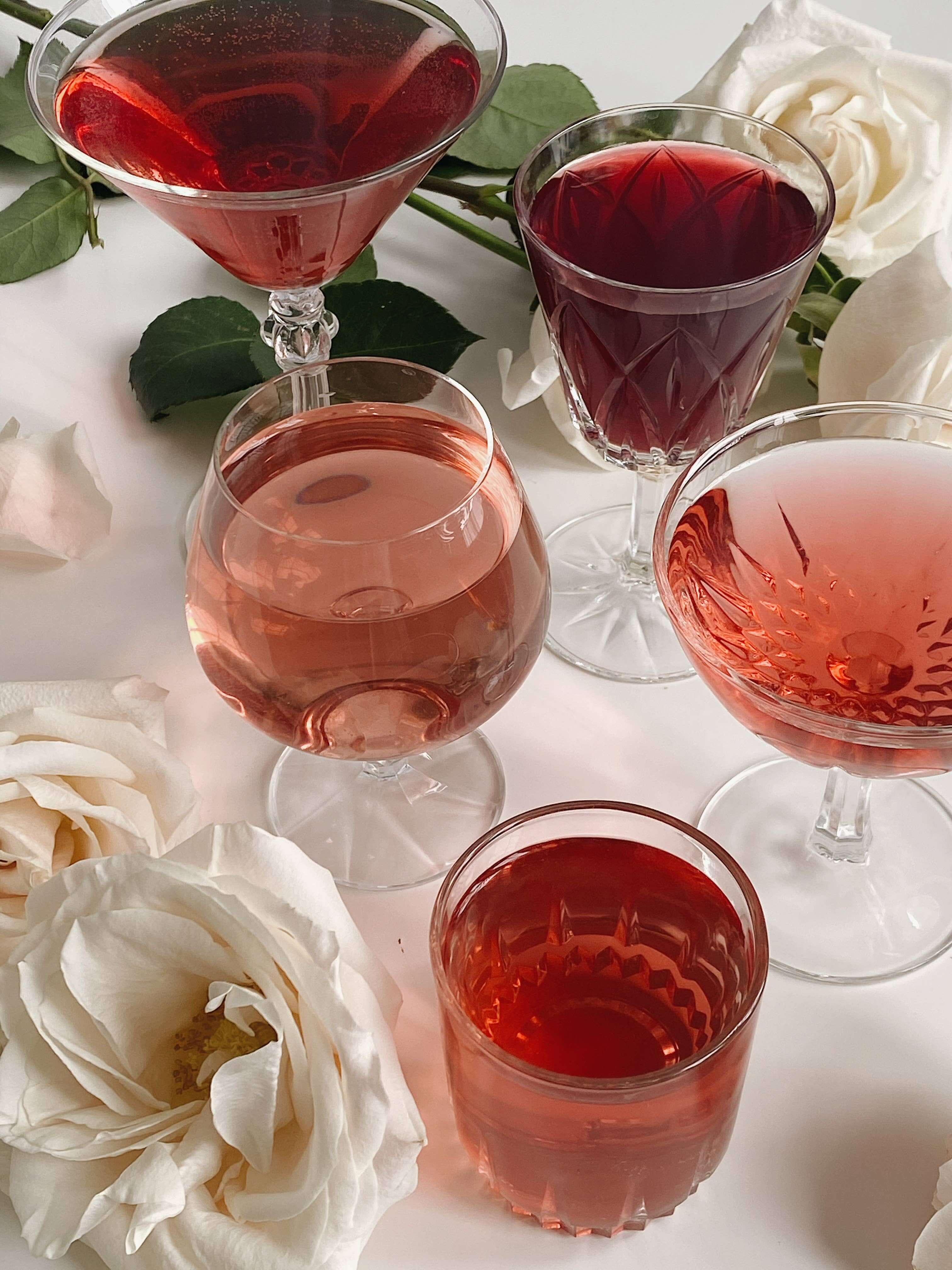 Assorted glasses of vibrant hibiscus tea drinks with white roses on a table, showcasing rose and fruity colors.