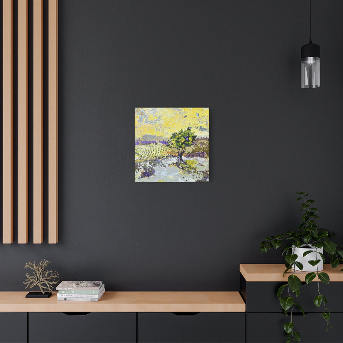 A small square canvas painting of a tree in a landscape hangs on a dark charcoal wall above a light wood console table in a modern room setting.