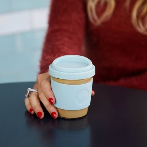 Hand holding an 8OZ Cool Cyan Glass Travel Tumbler by SOL Cups on a table, showcasing eco-friendly design.