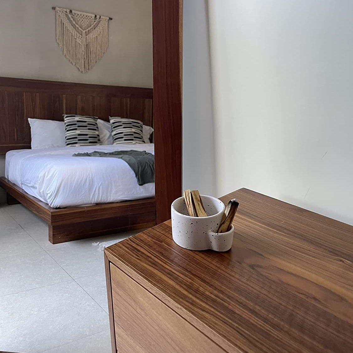 Wooden dresser with a mirror reflecting a bedroom with a bed and decorative wall hanging.